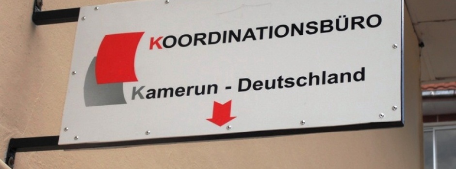 Das Bild zeigt ein Hausschild, auf dem das rot-weiße Logo des kamerunischen Alumnidachverband KBK zu sehen ist